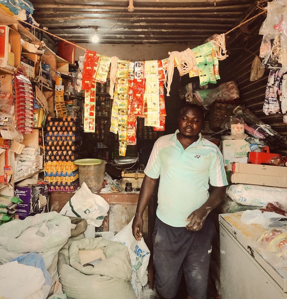 A shopkeeper standing in his duka, among sacks, egg trays and packets hanging from the roof
