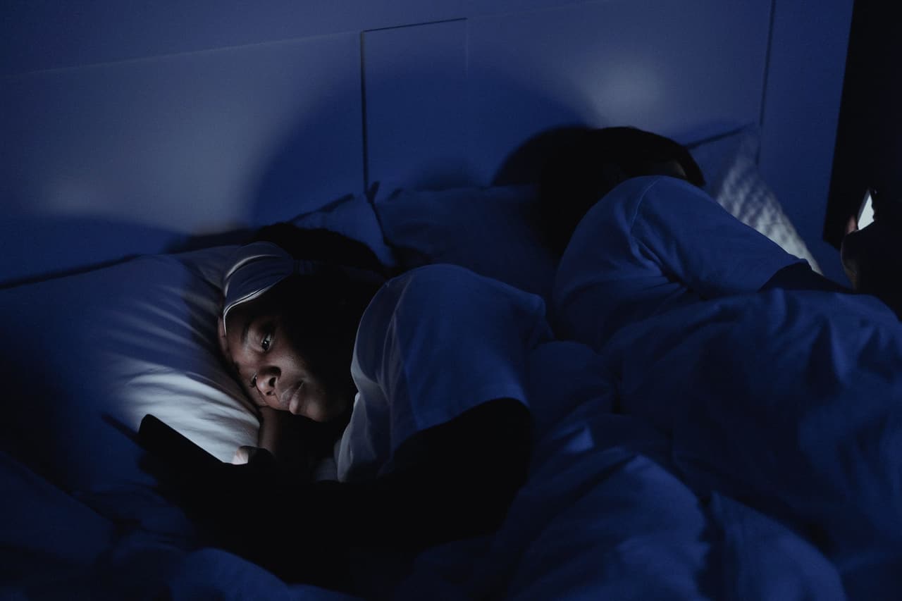 Night. One person asleep; beside them, a face lit by a phone screen