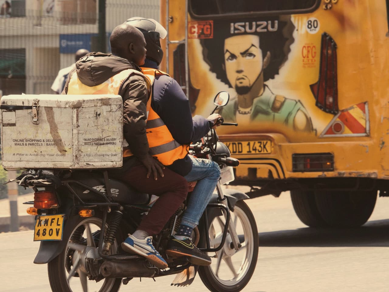 A Nairobi street in the evening: a boda boda with a delivery box passes a painted matatu